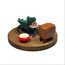 Sculpture of a child in a green kaiju costume laying on a braided carpet and watching a TV, with a bowl of popcorn and a toy robot figurine nearby. A monster movie is on the TV.