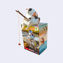 Small plastic cat sitting atop of a box, holding a branch with a fishing line attached. 
