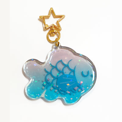Die cut semi transparent keychain of a blue fish with a closed eye smiling expression and glitter details. It is attached to gold star shaped hardware and has moving glitter within.