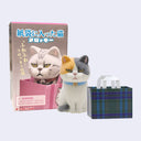 A toy cat figurine with a blue collar stands beside its pink retail box and a small plaid gift bag on a plain light background. The box shows a cat sitting in a pink paper bag with Japanese text and a small brand mark.