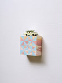 Small square wood panel painted like an orange-pink sunset with blue patterning overlayed on top, akin to a quilt tile design. Atop of it is a painted toy car with a wooden flower on top of its roof.