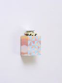 Small square wood panel painted like an orange-pink sunset with blue patterning overlayed on top, akin to a quilt tile design. Atop of it is a painted toy car with a wooden flower on top of its roof.