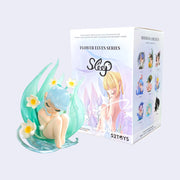 Vinyl figure of a nude, sleeping fairy with blue semi transparent hair with white flowers coming out of it. She sleeps within a teal colored leave that looks like an ocean wave. She sits next to her product packaging.