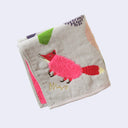 Light gray square hand towel with an illustration of stylized green and purple trees, with a fluffy pink fox standing under them. Close up to show embroidered details