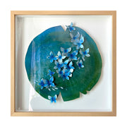 Painting on cut out paper of a round lily pad, blue and green with a series of small blue butterflies all over the pad like small flowers in a line. Piece is framed in thin, light grain wooden frame.