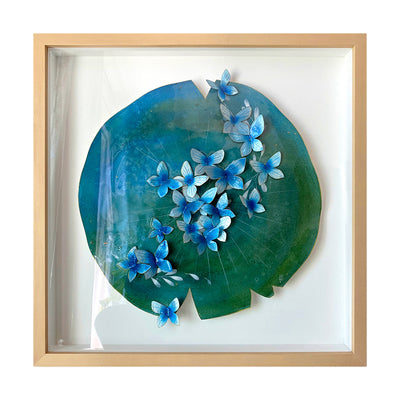 Painting on cut out paper of a round lily pad, blue and green with a series of small blue butterflies all over the pad like small flowers in a line. Piece is framed in thin, light grain wooden frame.