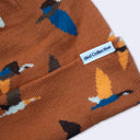 Close-up of a knit beanie in rust brown with a repeating bird pattern in blue, orange, white, and black. A white fabric label reading Bird Collective is sewn onto the cuff.