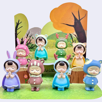 8 figurines of characters with calm, closed eye expressions wearing colorful cloaks with animal ears on the hoods. 