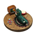 Sculpture of a child in a green kaiju costume laying on a braided carpet and watching a TV, with a bowl of popcorn and a toy robot figurine nearby. A monster movie is on the TV.