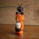 Painted whittled wooden sculpture of a fox with a surprised look on its face. It stands on its hind legs with its hand together. Atop its head is a small butterfly.