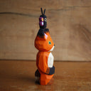 Painted whittled wooden sculpture of a fox with a surprised look on its face. It stands on its hind legs with its hand together. Atop its head is a small butterfly.