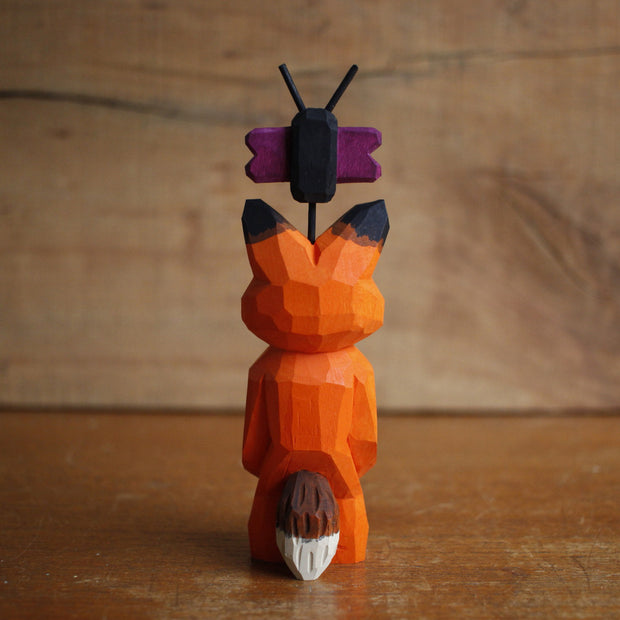 Painted whittled wooden sculpture of a fox with a surprised look on its face. It stands on its hind legs with its hand together. Atop its head is a small butterfly.