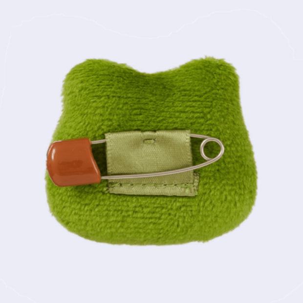 Green fabric plushie of a frog's head, with a safety pin on a light gray background