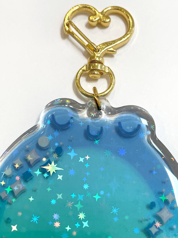 Die cut semi transparent keychain of a chubby blue frog, with its eyes closed and sparkles along its cheeks. It is attached to gold heart shaped hardware and has glitter within. Close up shot to show glitter.