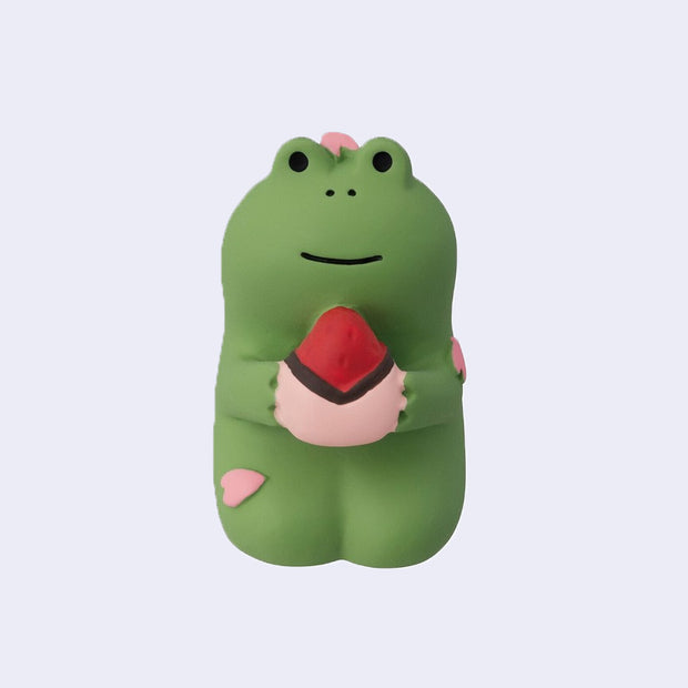 Small figure of a green frog sitting on its knees and holding a daifuku with cherry blossom petals atop its head and body.