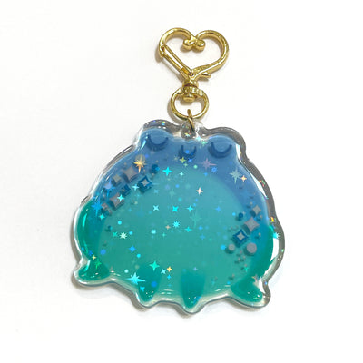 Die cut semi transparent keychain of a chubby blue frog, with its eyes closed and sparkles along its cheeks. It is attached to gold hardware and has glitter within.