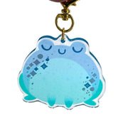 Die cut semi transparent keychain of a chubby blue frog, with its eyes closed and sparkles along its cheeks. It is attached to gold hardware and has glitter within.