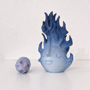A small wooden sculpture of a purple skull and a taller sculpture of a blue flame with a closed eyed simplistic face.
