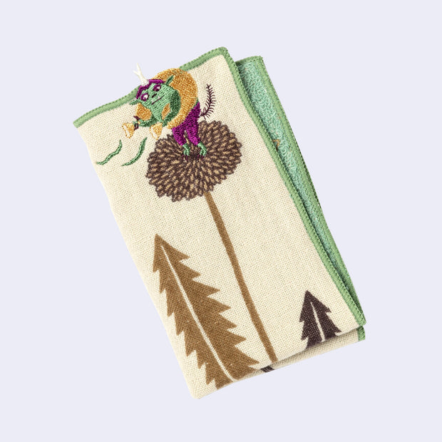 Rectangular cream colored towel with a green border, of a dandelion flower and pointed leaves. A small green yokai stands on top of the dandelion, with a bag of winds. Close up to show embroidered details.