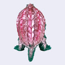 A toy monster figure of Gamera with a green body, pink shell, and yellow eyes, shown against a plain light gray background.