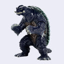 A small monster toy figure with a dark blue-black textured body, green shell, and white tusks, standing against a plain light gray background.