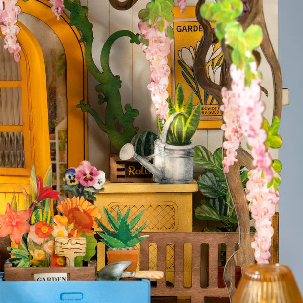 Detail shot of Holiday Garden House bookend diorama, featuring a tin watering can and vines of pink wisteria and flowers to be planted.