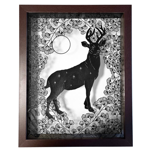  Black paper cutting in a shadowbox style frame with a glass back. A stag stands tall, with tiny stars on its body, around many intricately cut flowers and a floating circle. 