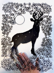 Black paper cutting art. A stag stands tall, with tiny stars on its body, around many intricately cut flowers and a floating circle. Held in the hand of the artist.