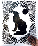 Black paper cutting art. A wolf sits and holds a star in its mouth, it is framed by delicately cut fern leaves and flowers and a full moon. It is held in the hand of the artist.
