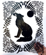 Black paper cutting art. A wolf sits and holds a star in its mouth, it is framed by delicately cut fern leaves and flowers and a full moon. It is held in the hand of the artist.