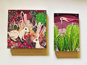 Set of 2 illustrative style paintings, both desert themed and featuring cacti. One is of 2 desert hares amongst cacti and the other is of a pink bird atop of round cacti.