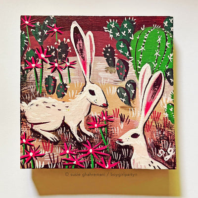 Illustrative style painting of 2 cream colored desert hares with long ears, standing in a desert setting with cacti and flowers around. Background is a reddish purple sky and brown flooring.