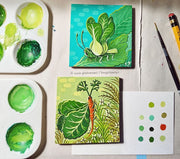 An artist's work station, with a paint brush, pencil, paint tray and a paper for color swatches. 2 artworks sit on the table, "Carrot Bug" and "Bok Choy Bug."
