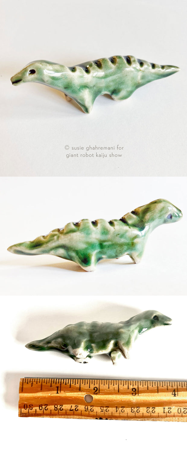 Small ceramic sculpture of a bluish green dinosaur with ridges along its back and pointed legs.