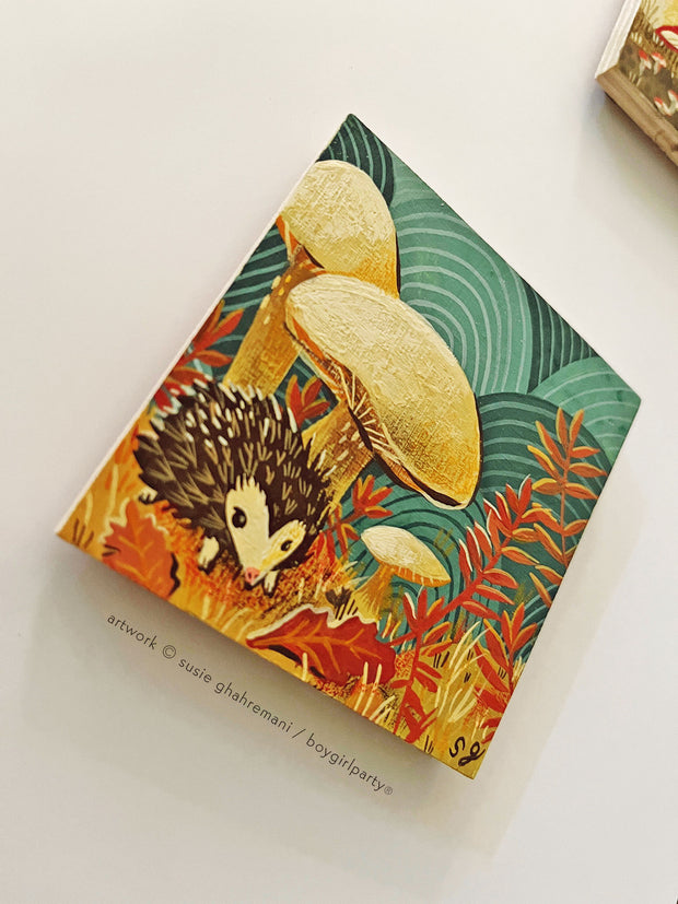 Painting of a small hedgehog under large yellow mushrooms. Ground is covered in autumn leaves and grass. Background is a pattern of teal concentric circles.