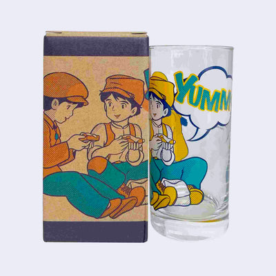 Glass cup with a screen printed design on the outside, imagery from Castle in the Sky of a girl and a boy, dressed in mechanic's working wear, sharing a meal of a slice of egg on toast. Large stylized text reads "YUMMY!!"