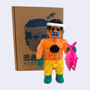 Colorful soft vinyl figure of an orange glittery man holding a pink fish in front of a cardboard box with a face design.