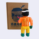 Colorful soft vinyl figure of an orange glittery man holding a pink fish in front of a cardboard box with a face design.