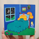 Painting held in the hand of the artist of a yellow cartoon character laying in a bed in a children's room. It has framed photos on the wall, small toy cars and a night sky visible from a nearby window.
