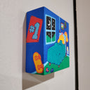 Painting held in the hand of the artist of a yellow cartoon character laying in a bed in a children's room. It has framed photos on the wall, small toy cars and a night sky visible from a nearby window. Shown at side angle.