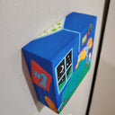 Painting held in the hand of the artist of a yellow cartoon character laying in a bed in a children's room. It has framed photos on the wall, small toy cars and a night sky visible from a nearby window. Shown from overhead.