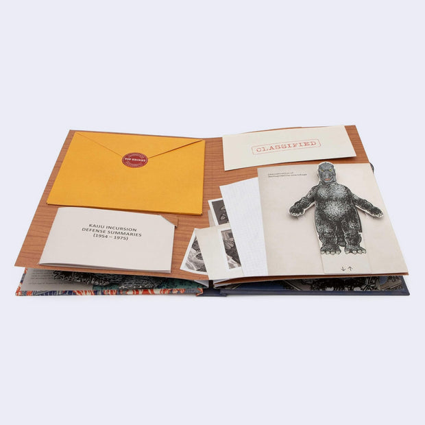 Open page from a pop up book about Godzilla, featuring a paper sculpture (pop up element) of Godzilla along with papers in various folders, that can be removed.