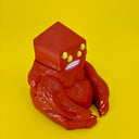 A wooden sculpture of a carved red demon, sitting on the ground. Its body is akin to a gorilla, and its head is pyramidal with 4 yellow eyes and straight white teeth.