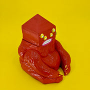 A wooden sculpture of a carved red demon, sitting on the ground. Its body is akin to a gorilla, and its head is pyramidal with 4 yellow eyes and straight white teeth.