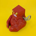 A wooden sculpture of a carved red demon, sitting on the ground. Its body is akin to a gorilla, and its head is pyramidal with 4 yellow eyes and straight white teeth. A wooden lever comes out of its side.