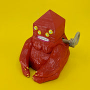 A wooden sculpture of a carved red demon, sitting on the ground. Its body is akin to a gorilla, and its head is pyramidal with 4 yellow eyes and straight white teeth. A wooden lever comes out of its side.