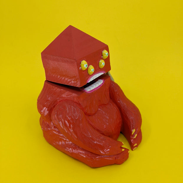 A wooden sculpture of a carved red demon, sitting on the ground. Its body is akin to a gorilla, and its head is pyramidal with 4 yellow eyes and straight white teeth. Its mouth is slightly ajar.