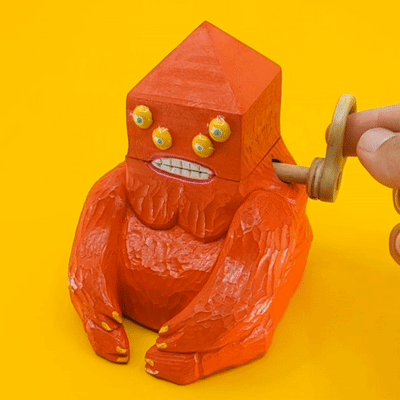 Gif of a wooden sculpture, a red demon with a triangular head and 4 yellow eyes. Its mouth opens and closes as a lever is turned.