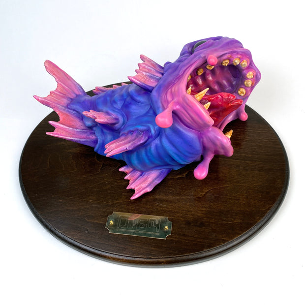 Pink and purple fish mounted to a wooden plaque. Its mouth is wide open, showing gold teeth and a red split tongue. Fish is hyper realistic in style.