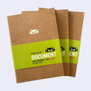 Three brown kraft paper sketchbooks with a bright green paper band around the middle, featuring a cartoon monster face logo and the text 'DOCUMENT KEEP DRAWING - 3PK BLANK'.